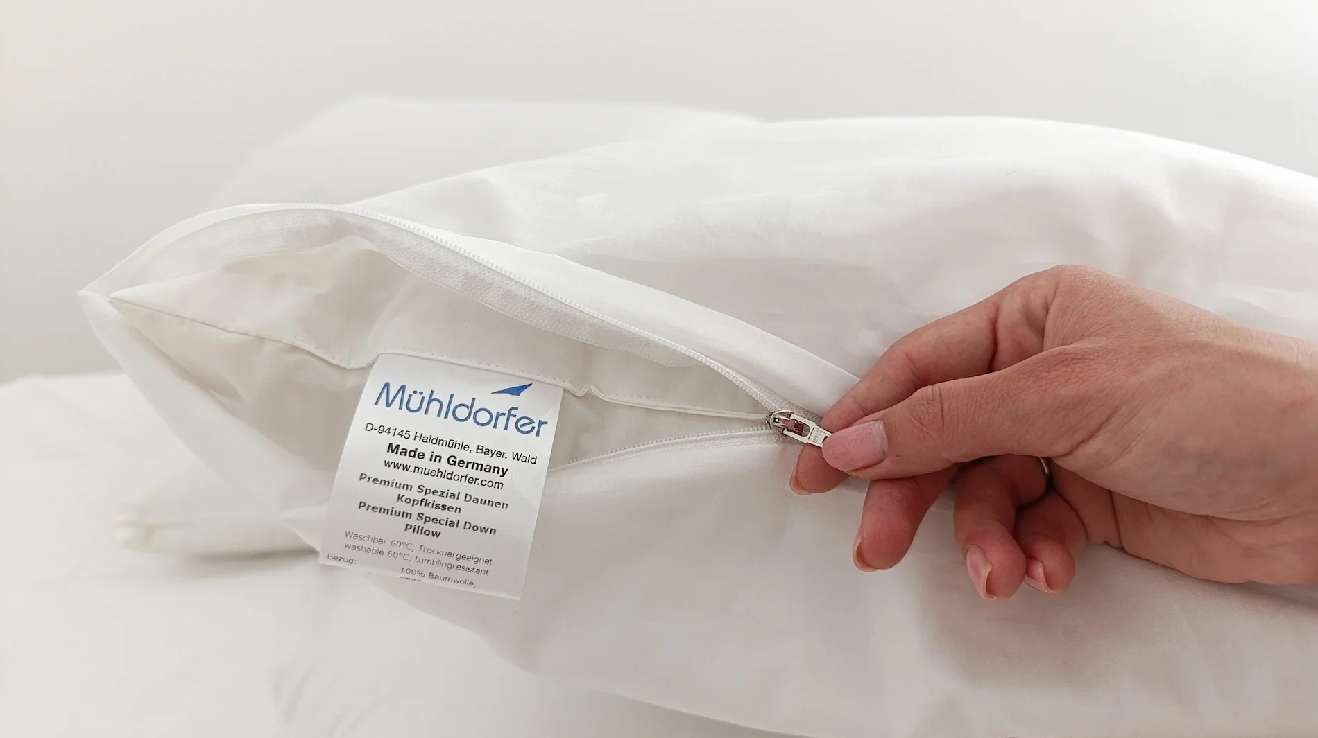 Hand opening white zippered pillow cover with Mühldorfer brand label showing quality details