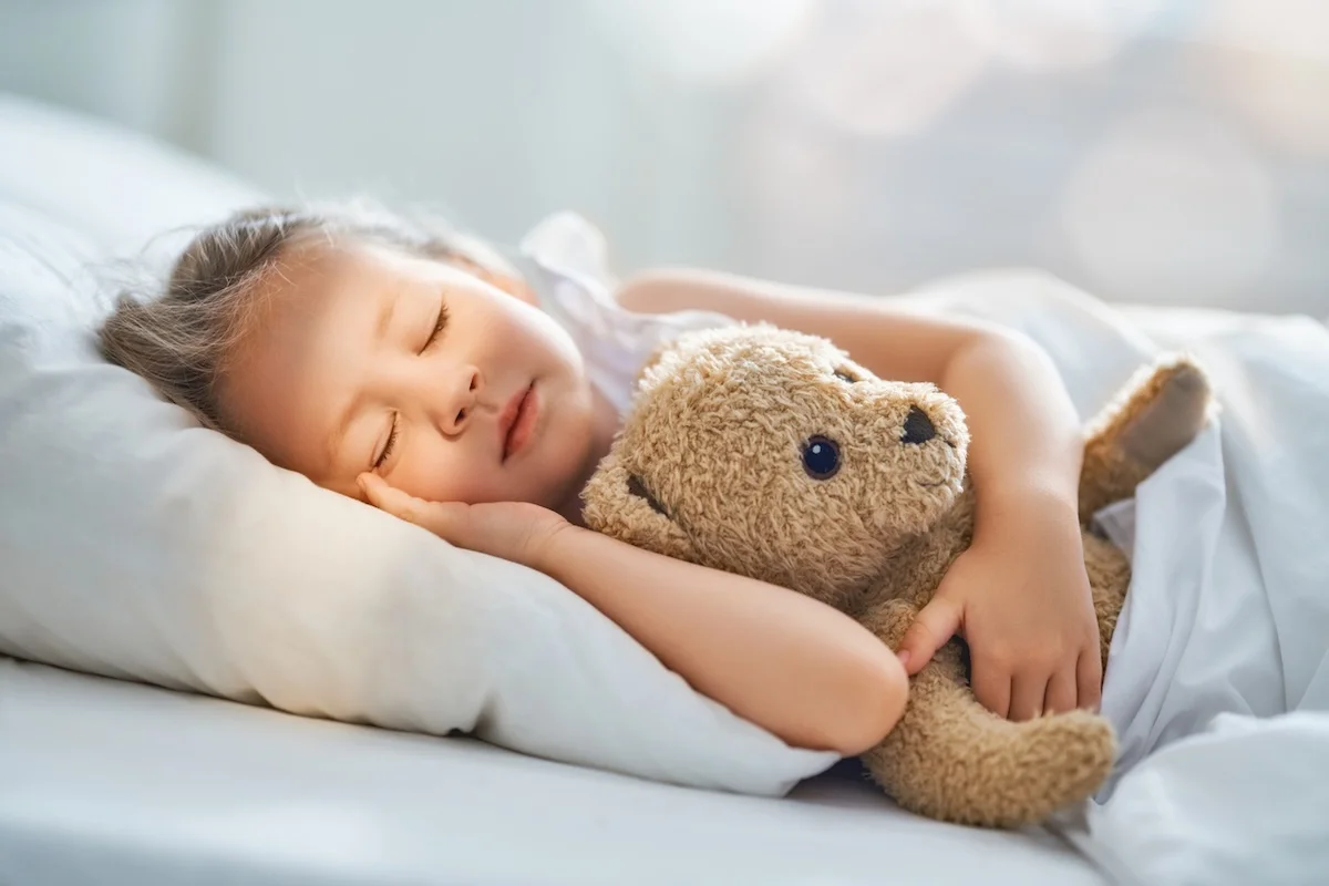Child sleeping peacefully under white kids duvet while cuddling brown teddy bear