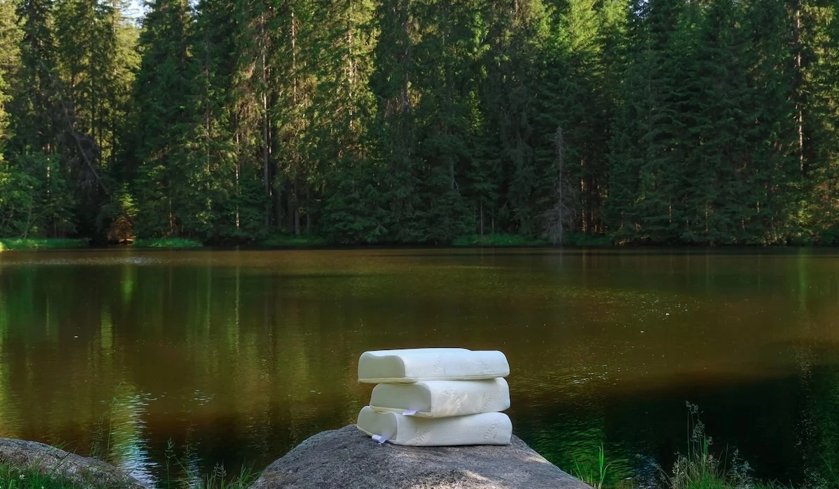 Three white ergonomic neck pillows stacked on rock beside forest lake
